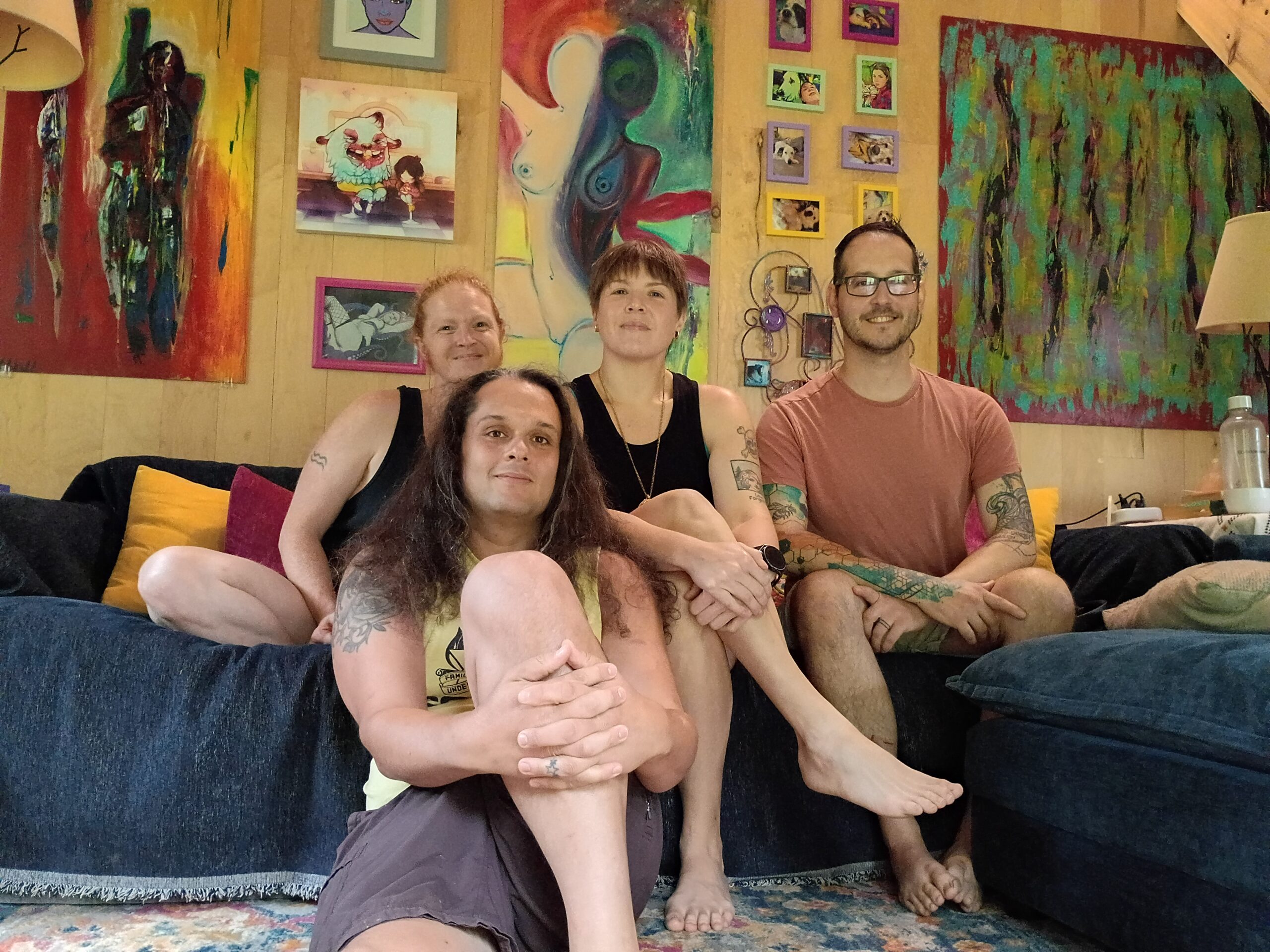 A group photo of four people, taken on a blue couch with lots of abstract art and photography hanging on a wall in the background.